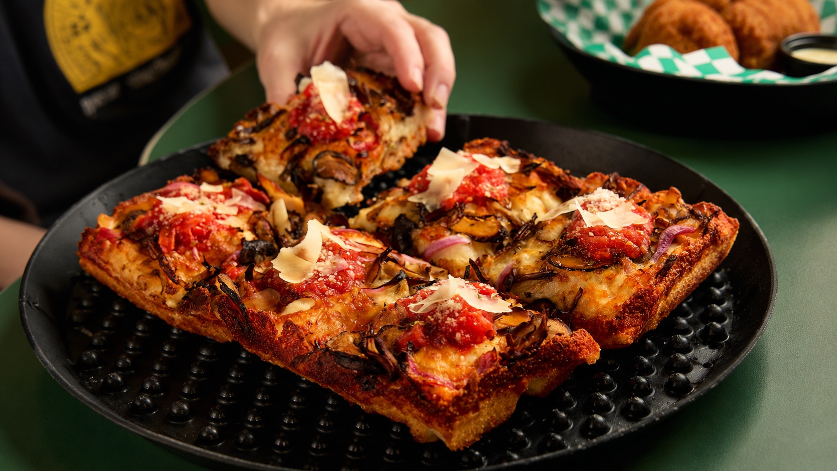 A hand lifting a square of Detroit-style pizza from a black plate