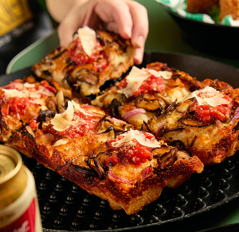 A hand reaching for a square slice from a Detroit-style pizza